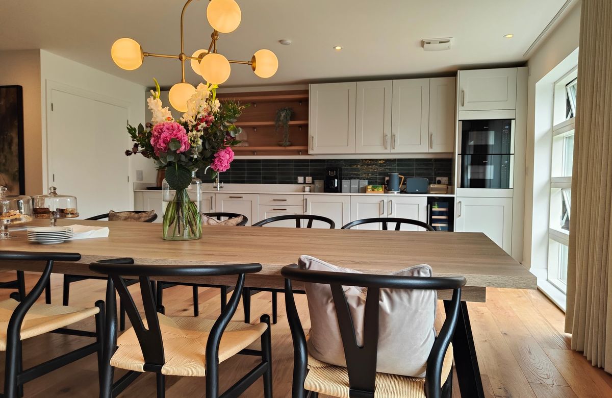 Kitchen cabinetry beyond a dining table with flowers and woven chairs.