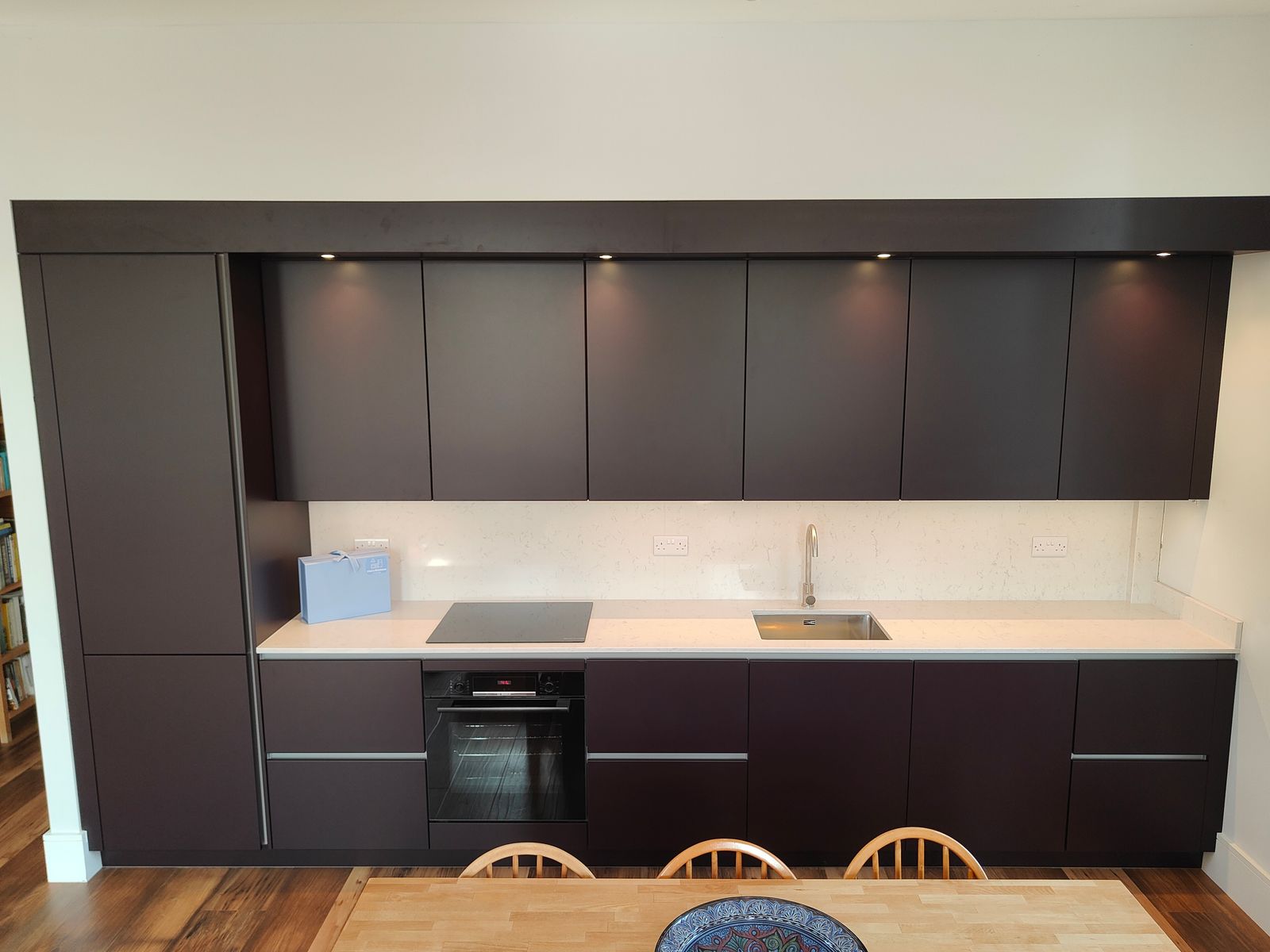 Front view of the aubergine kitchen run, with tall storage, an integrated oven, hob and sink behind the dining table.